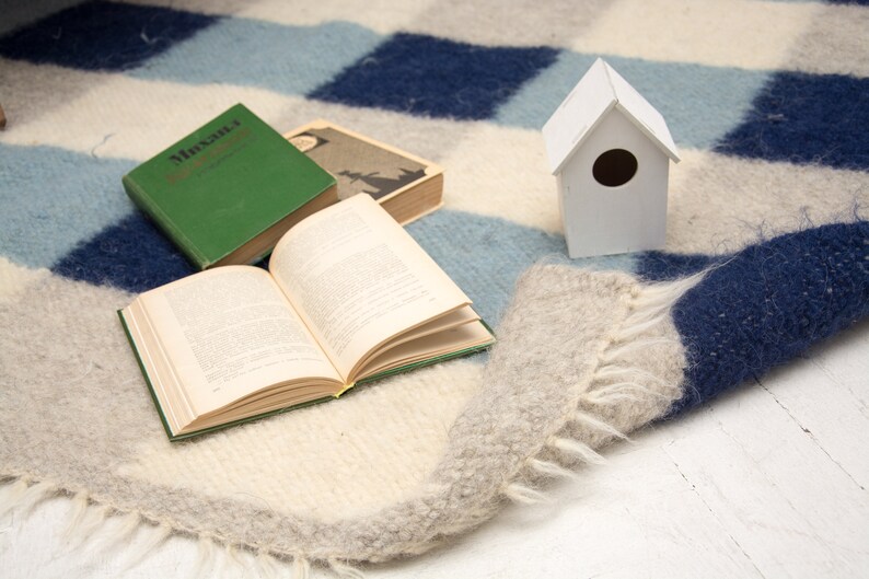 May include: A white birdhouse with a round opening sits on a blue and white striped rug with two open books. One book is green and has the title "Muscat" on the cover. The other book is open and has white pages.