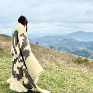 May include: A person standing on a grassy hilltop wearing a white and gray patterned blanket. The person is looking out at a distant mountain range.