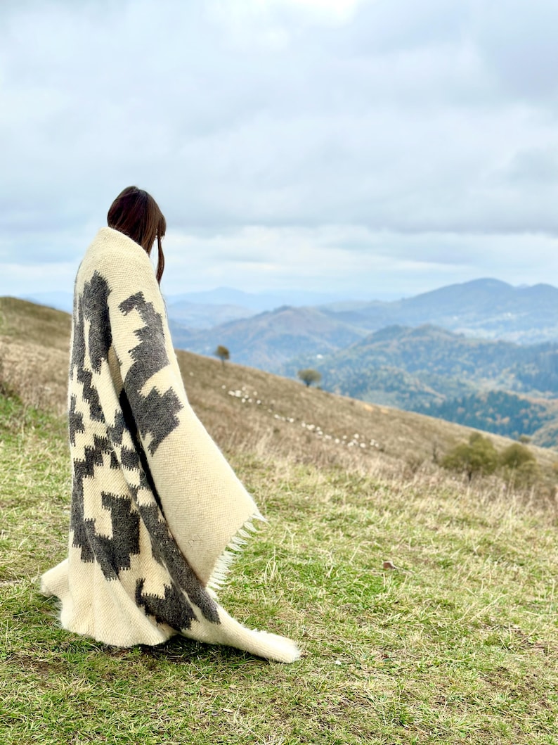 May include: A person standing on a grassy hilltop wearing a white and gray patterned blanket. The person is looking out at a distant mountain range.