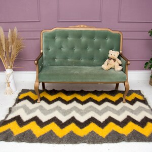 May include: A green velvet tufted sofa with a teddy bear sitting on the seat. The sofa is in front of a pink wall and a white floor. A gray and yellow chevron patterned rug is on the floor in front of the sofa.
