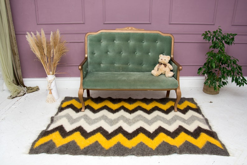 May include: A green velvet tufted sofa with a teddy bear sitting on the seat. The sofa is in front of a pink wall and a white floor. A gray and yellow chevron patterned rug is on the floor in front of the sofa.