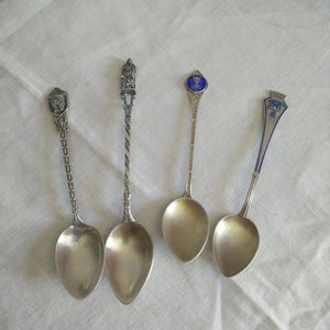May include: Four silver souvenir spoons with ornate handles. The spoons feature different designs, including a crown, a coat of arms, and a building.