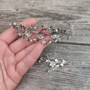 Może przedstawiać: Srebrna korona kwiatowa i pasująca spinka do włosów. Korona ma delikatne liście i kwiatowe detale z małymi, błyszczącymi akcentami. Spinka do włosów odzwierciedla wzór korony. Elementy są prezentowane na drewnianej powierzchni.