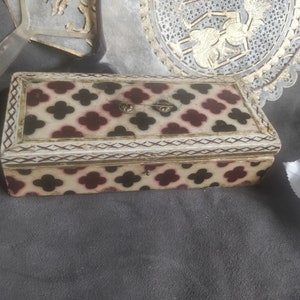 Può includere: Una piccola scatola rettangolare in legno con un coperchio incernierato. La scatola è decorata con un motivo ripetitivo di trifogli neri e rossi su sfondo bianco. Il coperchio ha un bordo dorato e due piccole cerniere metalliche.