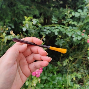 May include: A close-up of a dark purple and yellow plastic hair pick. The pick is held in a hand against a blurred background of green foliage.