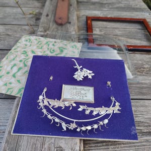 Puede incluir: Una corona floral de plata con un pequeño amuleto de flor de plata sobre un fondo de terciopelo azul. El amuleto tiene el texto "Gedenkrung 1945 Silberne Hochzeit".