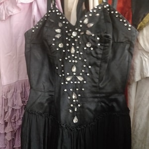May include: Black evening gown with a V-neckline and embellished with rhinestones. The dress has a fitted bodice and a flowing skirt. The straps are also adorned with rhinestones. Other dresses are visible in the background.