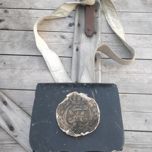 May include: A black leather bag with a white strap and a circular, decorative, paper patch on the front. The patch has a gold design.