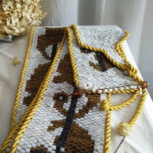 Könnte beinhalten: Ein weißer Perlengürtel mit einem braunen und schwarzen geometrischen Muster. Der Gürtel ist mit einem goldenen, seilartigen Besatz versehen.