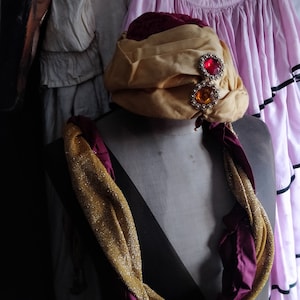 May include: A gold and burgundy turban with jeweled embellishments. The turban is on a mannequin with a gold and burgundy scarf.