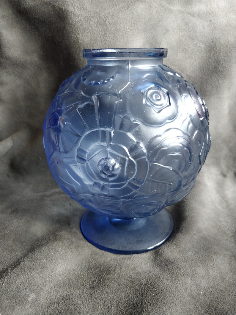 Op de afbeelding: Een blauwe glazen vaas met een rond lichaam en een brede basis. De vaas is versierd met een herhalend patroon van ge&euml;mbosseerde bloemen.