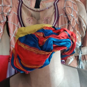 May include: A red, blue, and yellow turban with gold trim. The turban is made of fabric and is folded into a rounded shape.