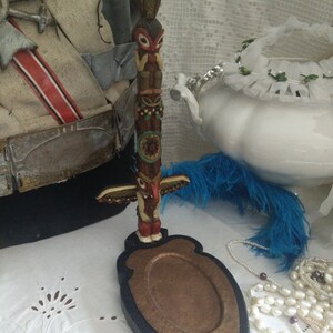 Puede incluir: Un tótem de madera con una base de color cobre. El tótem está pintado con diseños coloridos y presenta un pájaro, una cara y otras figuras. La base tiene forma de escudo y tiene un borde negro.