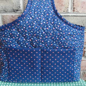 May include: A quilted tote bag in navy blue with a floral pattern of pink and white flowers. The bag has a large front pocket and a handle. The bag is designed for carrying items.