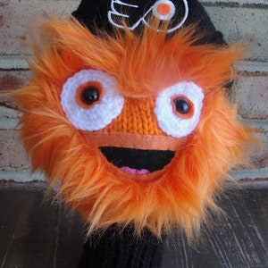 May include: A handmade orange and black Gritty-themed golf club head cover. The cover features a black knit base, orange fur, white and black eyes, and a smiling mouth. The top has the Philadelphia Flyers logo.