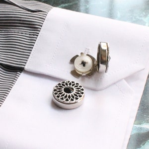 May include: A white dress shirt with a black and white striped pattern. The shirt has a white cuff with three silver cufflinks. Two of the cufflinks are round and have a black center. The third cufflink is a more intricate design with a black center.