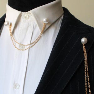 Op de afbeelding: Een wit overhemd met kraag onder een zwarte blazer met krijtstreep. De kraag is voorzien van een gouden dubbele ketting verbonden door twee witte kunstparels. Een bijpassende gouden broche met dubbele ketting en parel zit op de revers.