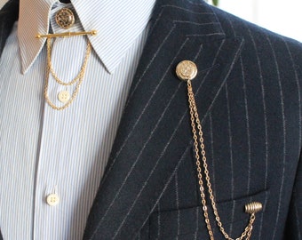 May include: A gold chain with a decorative button at the top is pinned to a dark blue pinstripe suit jacket. The chain is worn as a lapel pin.