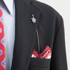 Puede incluir: Una chaqueta de traje negra con una camisa a rayas azules y blancas, una corbata de estampado rojo y un pañuelo de bolsillo de estampado rojo y blanco. Un broche de tortuga plateado está sujeto a la solapa.