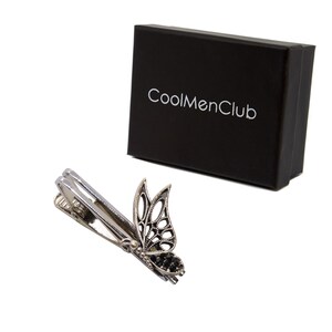May include: A silver-toned tie clip with a butterfly design, featuring openwork wings and black accents. The clip is next to a black box with the text "CoolMenClub" in white.
