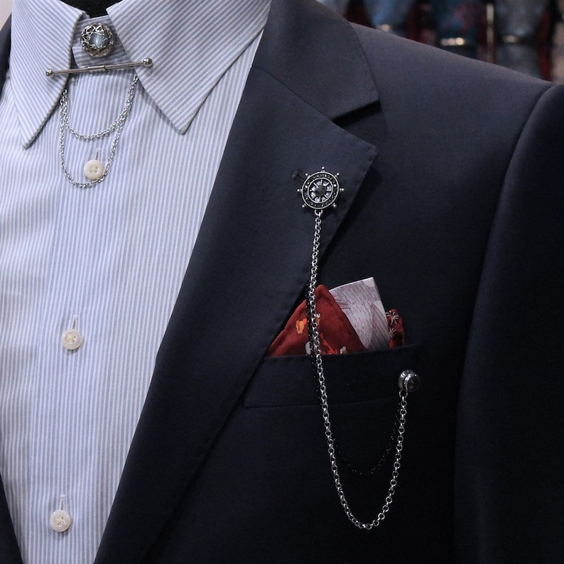 May include: A navy blue blazer with a pocket square and a silver chain with a decorative wheel charm. The chain is attached to a pin in the lapel.