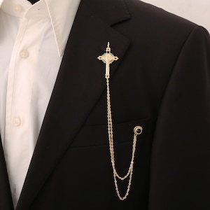 May include: A gold-toned cross pendant with a chain, worn as a lapel pin. The cross features a detailed design and a small black stone at the bottom of the chain.