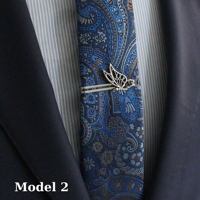 May include: A blue paisley patterned tie with a silver butterfly tie clip. The tie is worn with a navy blue suit jacket and a striped dress shirt. The text "Model 2" is visible in the bottom left corner.