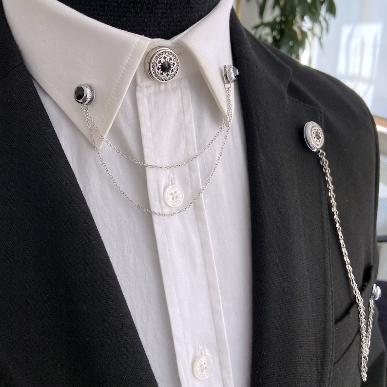May include: A white collared shirt with two silver chains attached to decorative buttons. The chains are connected to a black button on the collar and a silver button on the shirt.