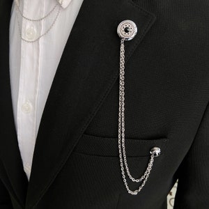 May include: A silver chain lapel pin with a black stone in the center. The pin is attached to a black suit jacket.