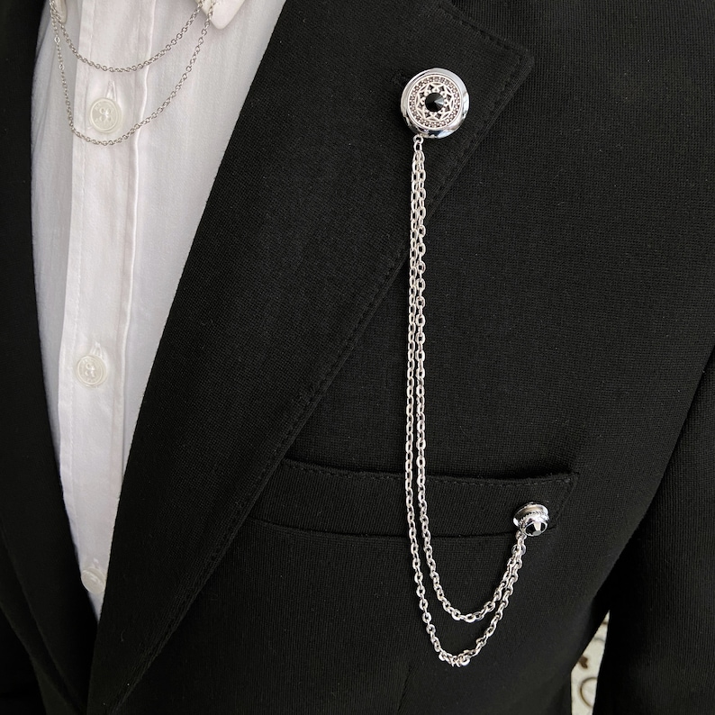 May include: A silver chain lapel pin with a black stone in the center. The pin is attached to a black suit jacket.