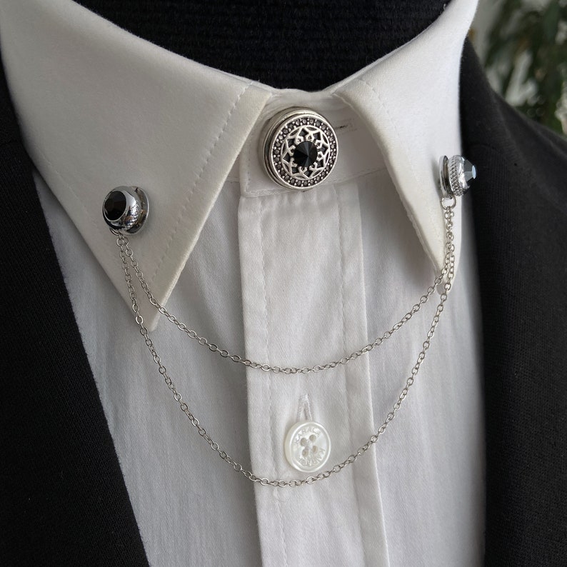 May include: A white collared shirt with a silver chain and two decorative collar pins. One pin features a black stone and the other has a silver and black design.