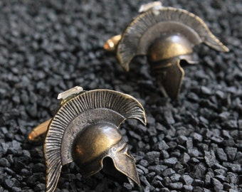 May include: A pair of gold-toned cufflinks featuring a detailed design of a Roman helmet. The helmets have a textured surface and a raised design.