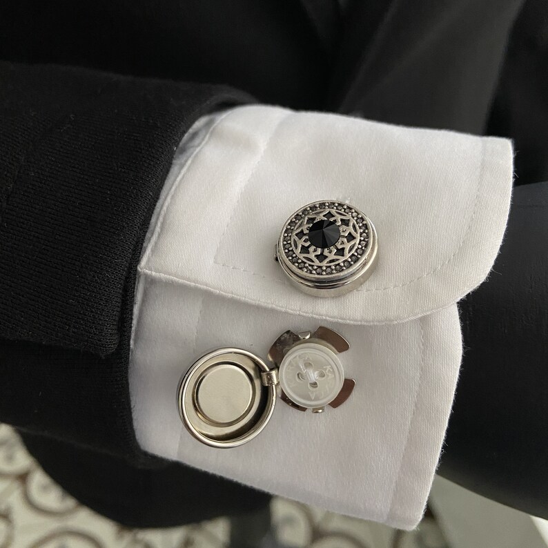 May include: A pair of cufflinks with a silver finish. One cufflink features a black stone surrounded by a decorative design. The other cufflink is a round, silver-toned button with a small, white button inside.