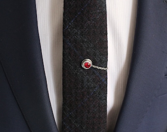 May include: A silver tie clip with a red gemstone. The tie clip is attached to a dark gray and blue patterned tie.