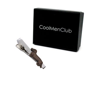 May include: A silver-toned tie clip with a brown dachshund dog design, next to a black box with the text "CoolMenClub" in white. The tie clip is angled, showing its clasp.