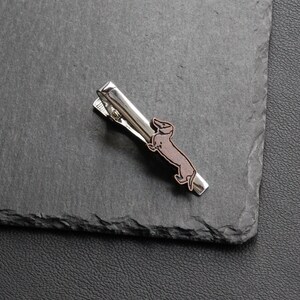 May include: A silver-toned tie clip featuring a brown and black dachshund dog design. The clip is rectangular with a hinged clasp and is set against a dark gray background.
