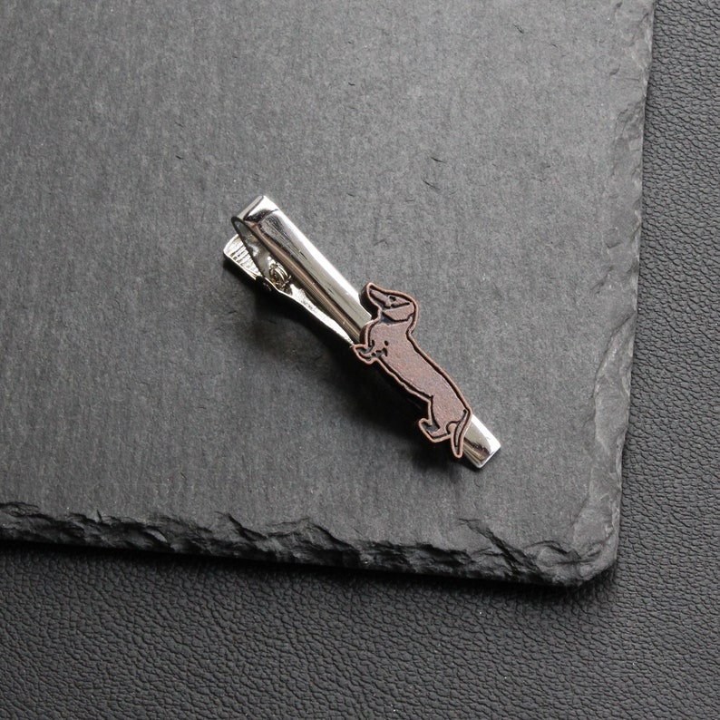 May include: A silver-toned tie clip featuring a brown and black dachshund dog design. The clip is rectangular with a hinged clasp and is set against a dark gray background.