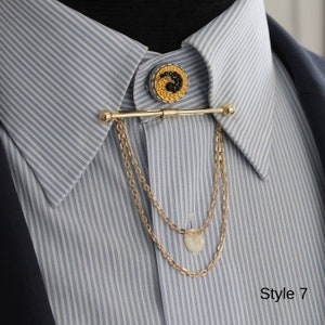 Pode incluir: Uma barra de colarinho dourada com uma corrente decorativa e um bot&atilde;o redondo. O bot&atilde;o apresenta um design preto e dourado. A barra de colarinho est&aacute; presa a uma camisa listrada azul e branca. O texto "Style 7" &eacute; vis&iacute;vel.