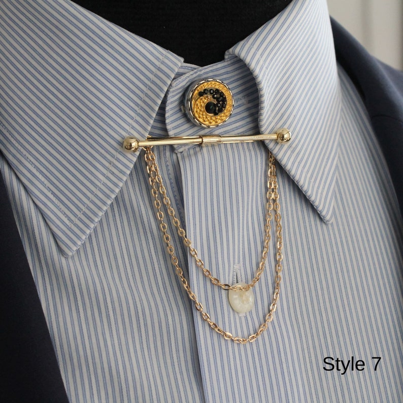 Pode incluir: Uma barra de colarinho dourada com uma corrente decorativa e um bot&atilde;o redondo. O bot&atilde;o apresenta um design preto e dourado. A barra de colarinho est&aacute; presa a uma camisa listrada azul e branca. O texto "Style 7" &eacute; vis&iacute;vel.