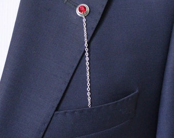 Puede incluir: Una cadena de plata con una joya roja unida a un broche de solapa. El broche está unido a una chaqueta de traje azul oscuro.