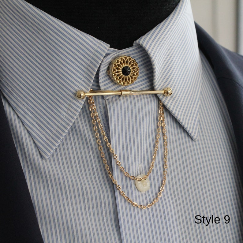 Pode incluir: Alfinete de colarinho dourado com um design floral decorativo e uma pedra central preta. O alfinete apresenta uma barra horizontal com correntes penduradas, adicionando um toque de eleg&acirc;ncia a uma camisa listrada azul e branca. O texto "Style 9" &eacute; vis&iacute;vel.