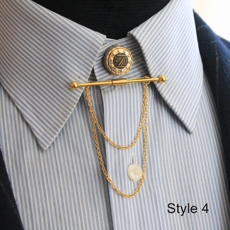 Pode incluir: Um alfinete de colarinho dourado com um bot&atilde;o decorativo e detalhe de corrente. O alfinete est&aacute; preso a uma camisa listrada azul clara e branca. O bot&atilde;o tem um design circular com um logotipo. O texto "Style 4" &eacute; vis&iacute;vel na imagem.