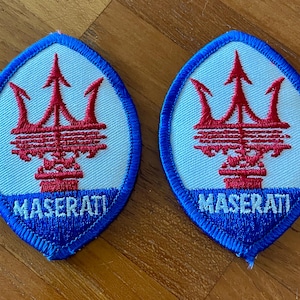 Puede incluir: Dos parches bordados de Maserati. Presentan un tridente rojo sobre fondo azul claro, con la palabra "MASERATI" en blanco. Los parches ovalados tienen un borde azul.
