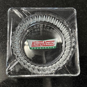 May include: A clear glass square ashtray with a raised, textured rim. The center of the ashtray features a green and red Krispy Kreme Doughnuts logo.