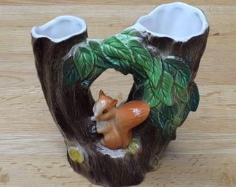 May include: A ceramic vase shaped like a tree trunk with two openings, decorated with green leaves and a brown squirrel figurine.