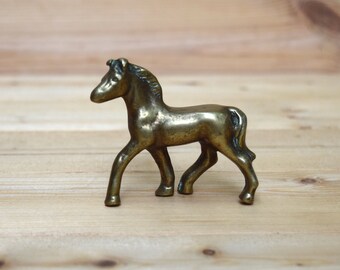 May include: A small, brass figurine of a horse standing on a wooden surface. The horse is facing to the left and has a detailed mane and tail.
