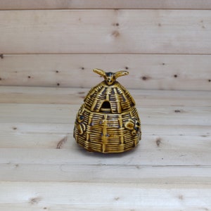 May include: A ceramic honey pot shaped like a beehive with a bee on top. The pot is brown and yellow with black accents.