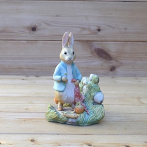 May include: A ceramic figurine of Peter Rabbit, a character from the children's book by Beatrix Potter. The figurine is painted in realistic colours and depicts Peter Rabbit standing on a grassy base with a red and white scarf.