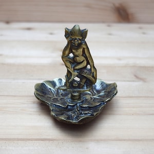 May include: A brass trinket dish with a detailed design of a flower. A small, detailed brass figurine of a goblin sits atop the dish.