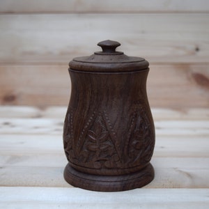 May include: A dark brown wooden jar with a lid, intricately carved with a floral pattern. The jar has a rounded base and a slightly flared top.
